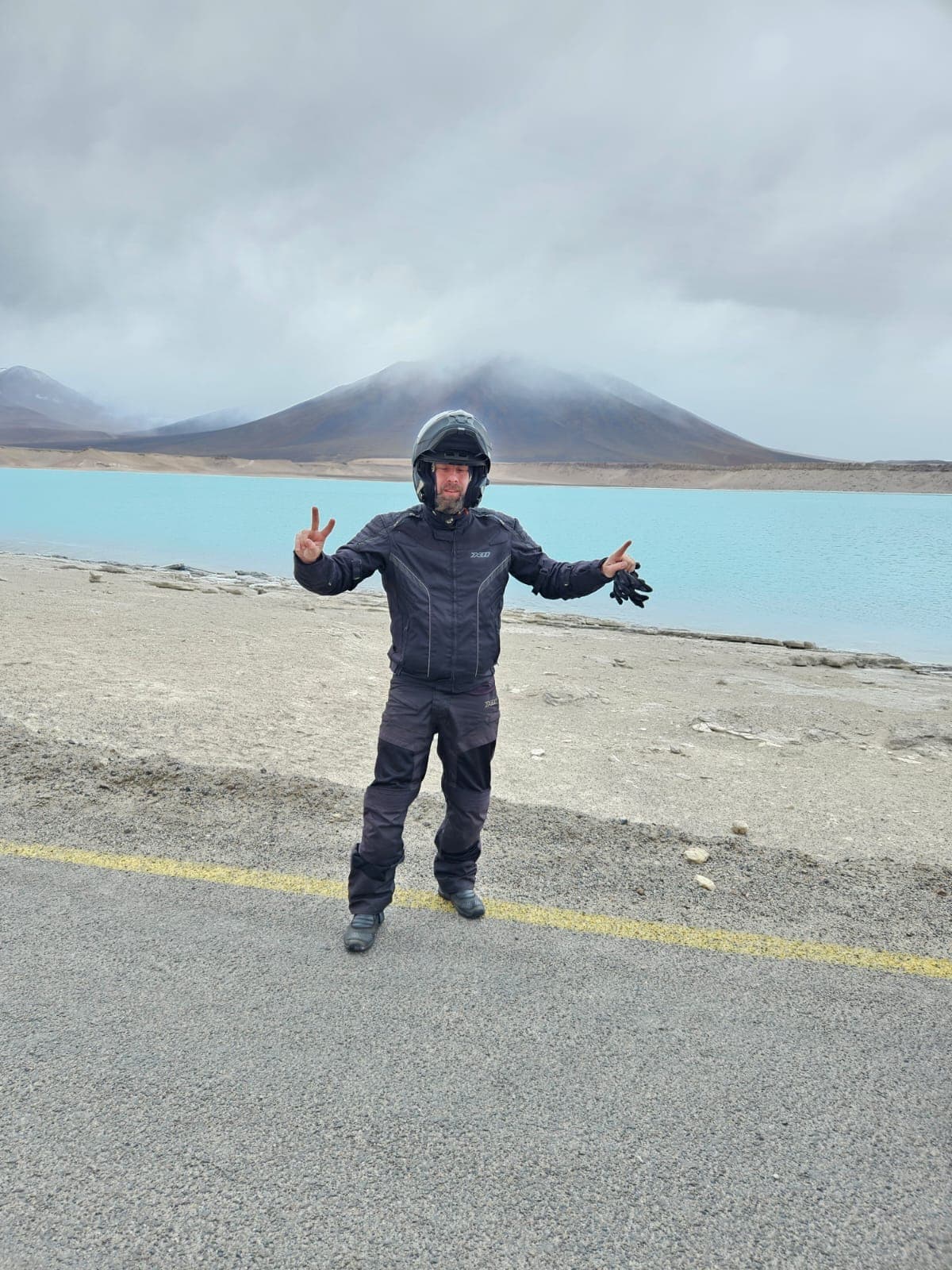 Motociclista de braços abertos na margem de uma lagoa turquesa, com montanhas ao fundo