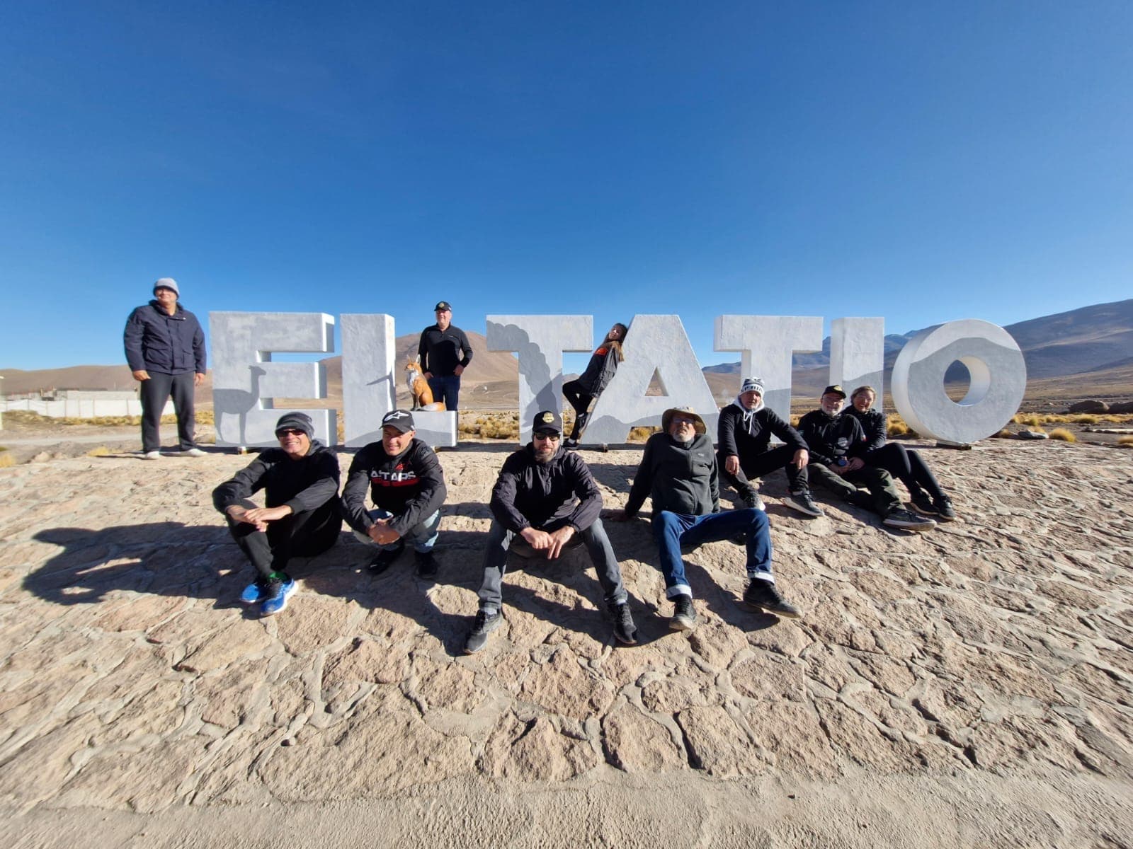 Grupo sentado na areia diante do letreiro de El Tatio, sob céu azul