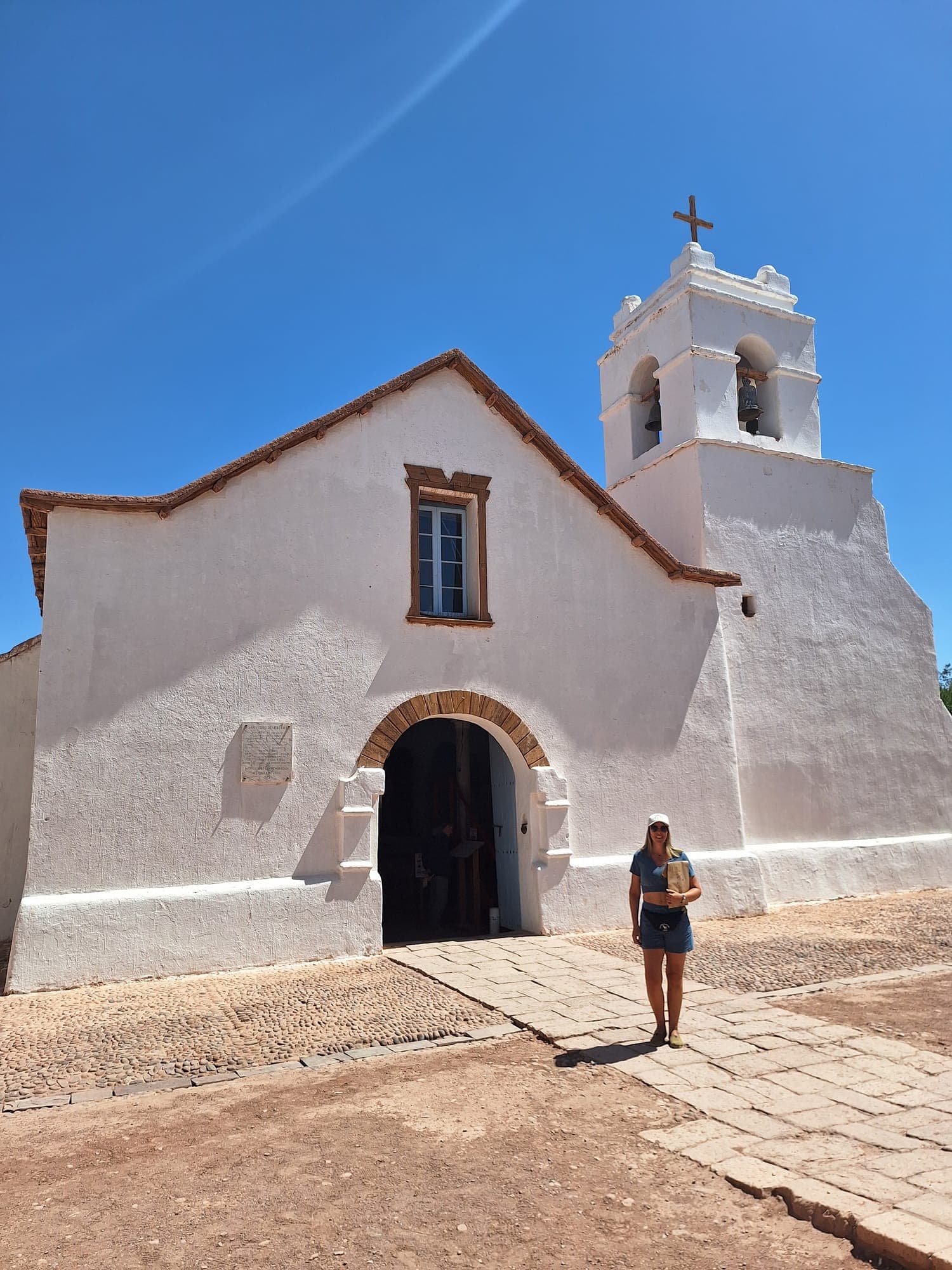 Fachada branca de uma igreja de adobe com torre e cruz, sob céu azul intenso