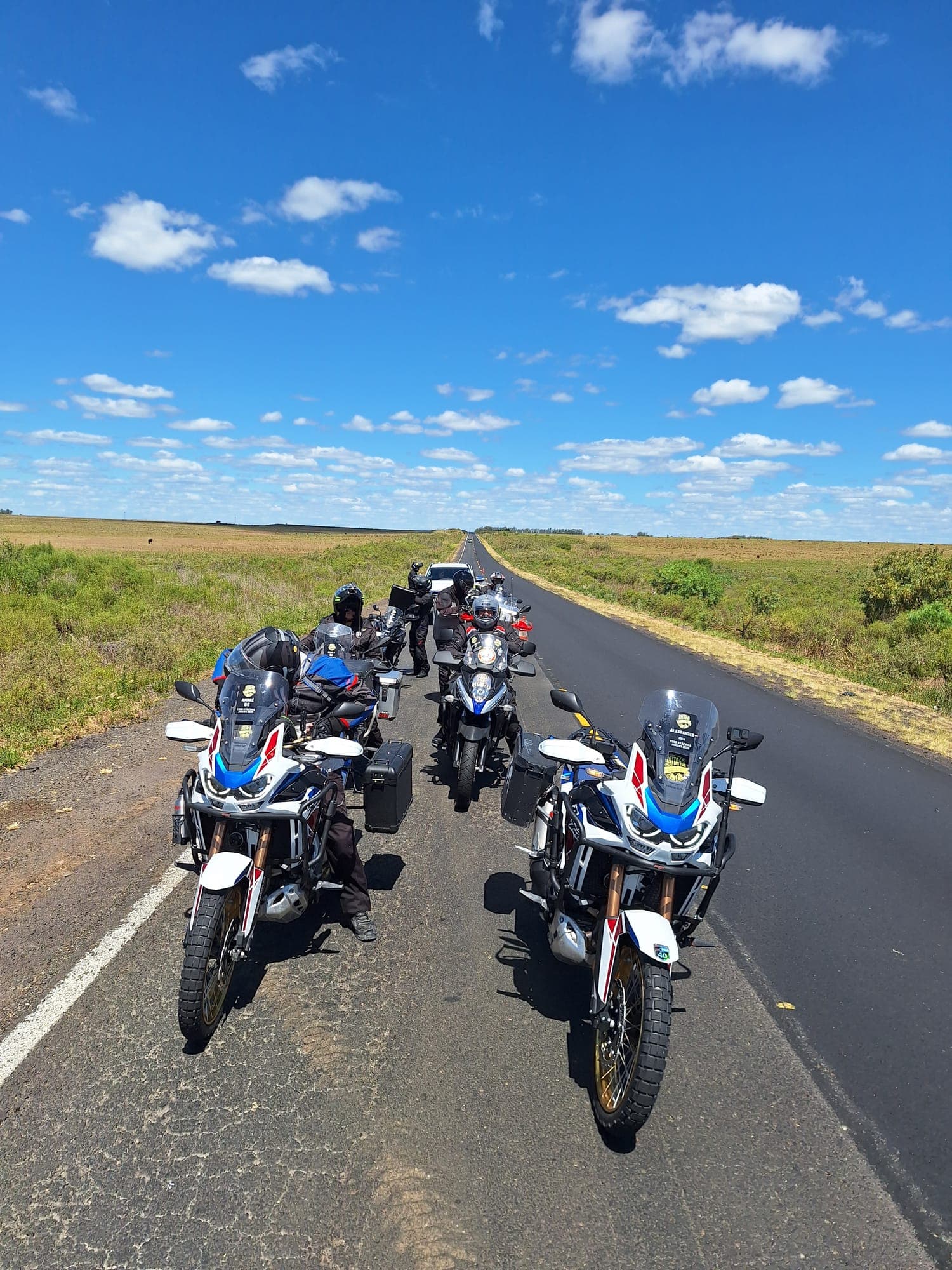Duas motos de aventura paradas na pista, com campo verde e céu azul