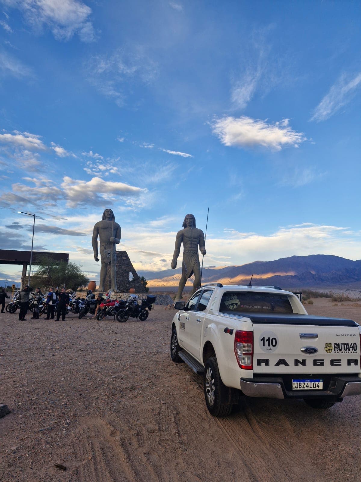 Caminhonete de apoio branca em primeiro plano, com as motos do grupo e as estátuas gigantes de Fiambalá ao fundo